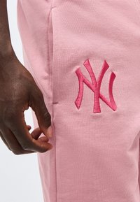 Dunkelhäutige Hand ruht in der Nähe einer Seitentasche einer hellrosa Hose mit einem dunkelrosa bestickten "NY"-Logo.