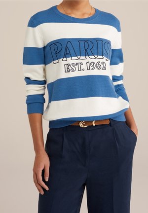 Personne portant un pull rayé bleu et blanc avec l'inscription "PARIS EST. 1962", un pantalon bleu marine et une ceinture marron avec une boucle dorée.