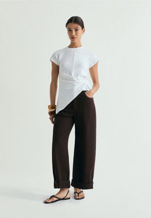 Donna in piedi che indossa un top bianco asimmetrico, pantaloni neri a gamba larga risvoltati, sandali neri e braccialetti in legno a un polso.