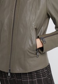 Oliivinvihreä nahkatakki, jossa on sileä pinta, vetoketjullinen etuosa ja sivutaskut. Varustettu hopeanvärisillä vetoketjuilla ja muotoillulla istuvuudella.