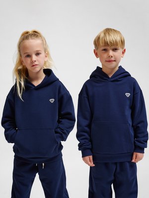 Twee kinderen dragen bijpassende marineblauwe hoodies en broeken en staan naast elkaar tegen een eenvoudige achtergrond.