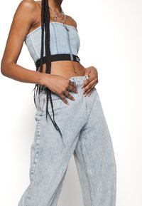 Top in denim corto con zip, di colore azzurro chiaro, abbinato a jeans blu chiaro a vestibilità morbida. Capelli intrecciati e una collana di perline multi-colore completano il look.