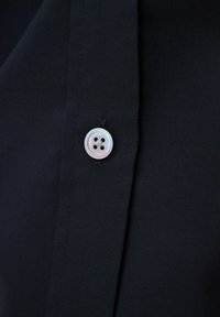 Czarna tkanina koszuli o gładkiej fakturze, z okrągłym, perłowym guzikiem z czterema otworami, przymocowanym przy listwie.