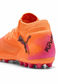 Scarpetta da calcio arancione brillante con collo in maglia, tomaia testurizzata, logo Puma nero e accenti rosa su tallone e suola, con molteplici tacchetti.
