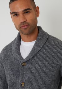 Szary dziergany kardigan z kołnierzem typu szalowego, z drewnianymi guzikami i prążkowaną fakturą. Noszony na białej koszulce T-shirt.