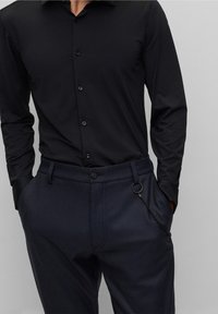 Camicia nera a maniche lunghe con chiusura a bottoni, abbinata a pantaloni scuri con motivo, dotati di tasca con cerniera e dettaglio ad anello metallico.