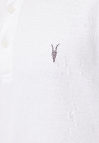 Witte poloshirt met een gestructureerd oppervlak, voorzien van een knoopsluiting en een klein, grijs geborduurd geitenkoplogo op de borst.