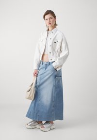 Bela denim jakna z žepi, v kombinaciji z svetlo modrim maxi denim krilom in srebrnimi natikači. Bež torba je preko ramena.