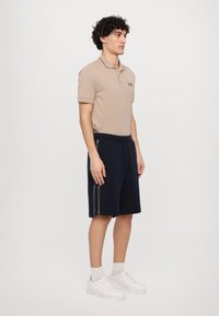 Béžová polo košile s krátkým rukávem a logem, kombinovaná s námořnickými šortkami se šedými bočními pruhy. Stylizováno s bílými teniskami.