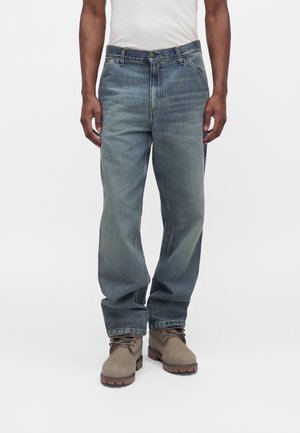 Mann trägt locker sitzende, ausgewaschene blaue Jeans, weißes T-Shirt und braune Schnürstiefel, steht vor schlichtem weißen Hintergrund.