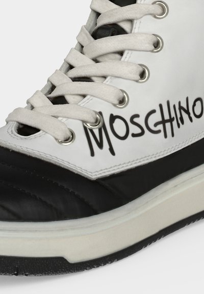 Gros plan d'une basket noir et blanc avec des lacets blancs et "Moschino" écrit en noir sur le côté au-dessus de la semelle.