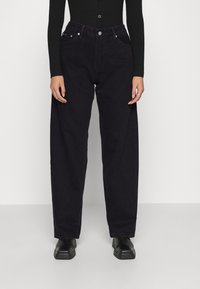 Persona vestida con jeans negros de tiro alto y corte ancho, una blusa negra de manga larga y botas negras de punta cuadrada, de pie frente a un fondo liso.