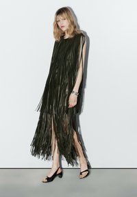 Vrouw met blond haar draagt een mouwloze zwarte franjesjurk en zwarte schoenen met open tenen, staand tegen een effen witte achtergrond.