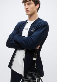 Giacca con zip blu navy con accenti bianchi, indossata sopra una maglietta bianca. Il modello tiene in mano una racchetta da tennis, con le braccia incrociate, con una texture liscia del tessuto.