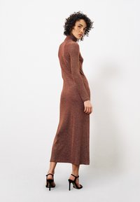 Langes, schlank geschnittenes Kleid aus schimmerndem Roststoff, mit einem hohen Kragen und langen Ärmeln, kombiniert mit schwarzen, schnürbaren High Heels.