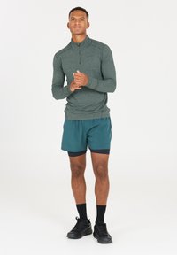 Langärmliges, mit Reißverschluss versehenes, dunkelgrünes Sportoberteil, kombiniert mit türkisfarbenen Shorts. Schwarze Sportschuhe und Socken runden das Outfit ab. Einfaches Design, keine Muster.
