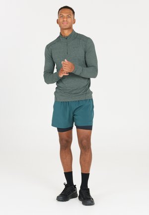 Mörkgrön träningsöverdel med lång ärm och dragkedja, som är ihopparad med petrolfärgade shorts. Svarta träningsskor och strumpor fulländar outfiten. Enkel design, utan mönster.