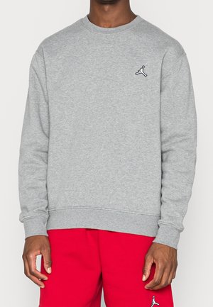 Osoba ubrana w szarą bluzę z długim rękawem i czerwone spodnie, obie z małym czarno-białym logo Jumpman.