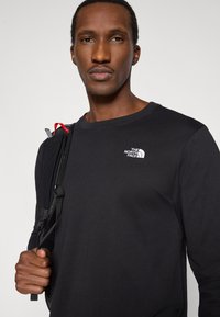 Μαύρο πουκάμισο με μακριά μανίκια και στρογγυλή λαιμόκοψη, με λευκό λογότυπο στο αριστερό μέρος του στήθους και υφή υφάσματος. Ορατό ρυθμιζόμενο λουρί από σακίδιο πλάτης.