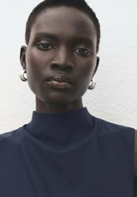 Granatowy bezrękawnik z wysokim kołnierzem. Gładka tekstura materiału. Modelka nosząca srebrne kolczyki w kształcie kół. Neutralne tło.