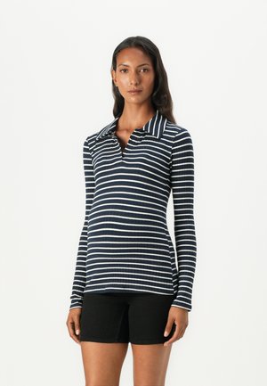 Marineblå og hvite stripete langermet topp med ribbet tekstur, med krage. Båret med svarte shorts, poserer modellen mot en hvit bakgrunn.