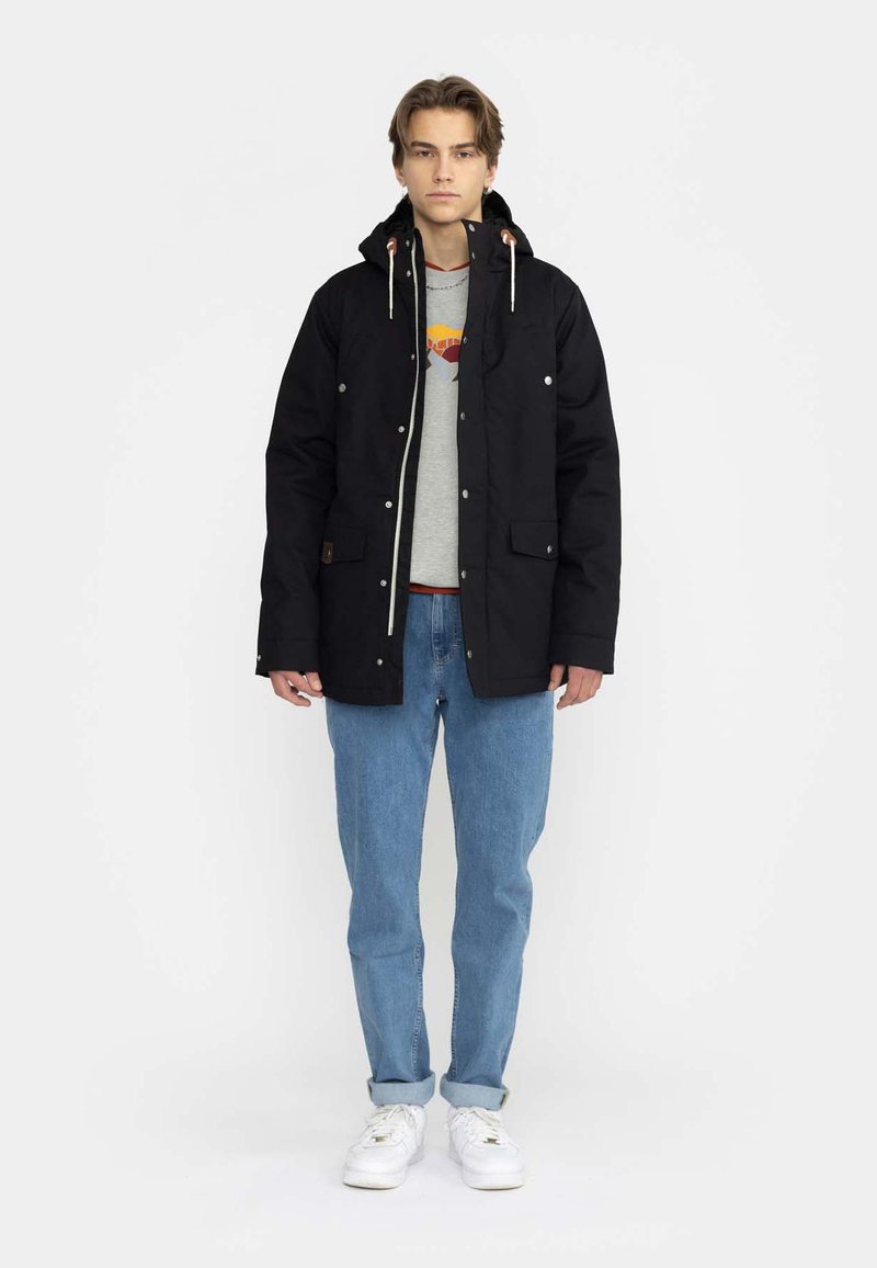 Parka nera con tasche frontali, cappuccio regolabile e accessori in argento, indossata sopra un maglione grigio e jeans blu. Sneakers bianche.