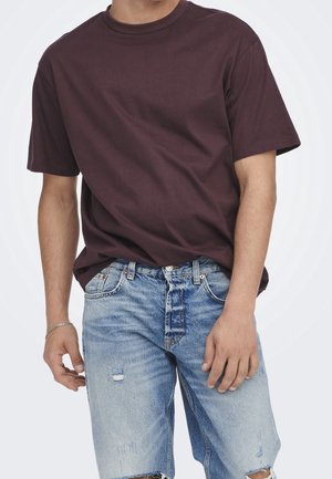 Homme portant un t-shirt uni marron foncé à manches courtes et un short en jean bleu clair déchiré avec un bracelet argenté au poignet gauche.