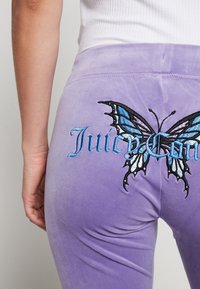 Lila sammetstights med en broderad blå fjäril och "Juicy"-text på baksidan. Slät textur, åtsittande design.