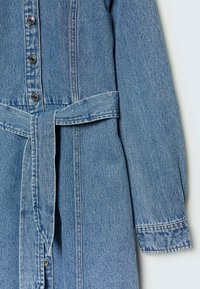 Denim-mekko, jossa on napit etuosassa, vyötärö on kireä sidotulla vyöllä ja pitkähihaiset hihat. Väri on vaaleansininen ja siinä on tekstuuripinta.