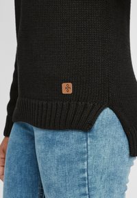Schwarzer Strickpullover mit geripptem Saum und seitlichen Schlitzen, versehen mit einem kleinen braunen Logo-Anhänger. Kombiniert mit hellblauen Jeans.