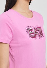 Růžové bavlněné tričko s krátkým rukávem a kulatým výstřihem, s lesklým logem "EA7" v stříbrné a růžové na přední straně.