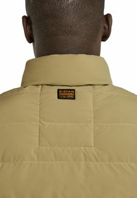 Chaqueta de plumas color caqui con cuello alto, que presenta una textura acolchada. La parte posterior muestra una etiqueta con el logo de G-Star sujeta en el cuello.