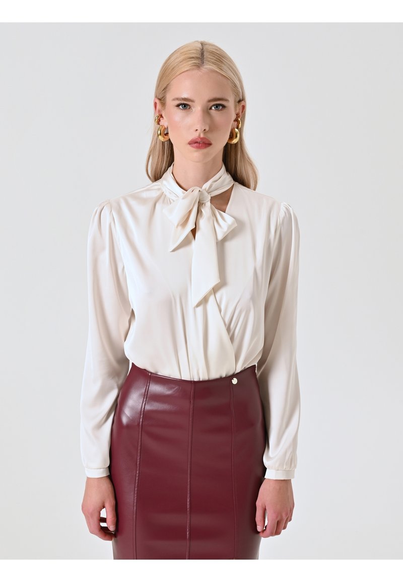 Blusa crema con un fiocco annodato al collo, maniche lunghe con spalle plissettate, abbinata a una gonna a matita in pelle bordeaux.