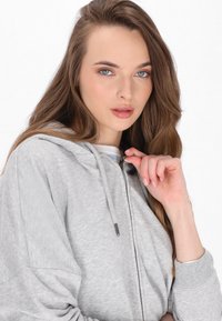 Šedá mikina na zip vyrobená z měkké strukturované látky s kapucí na stahovací šňůrku. Disponuje minimalistickým designem a žebrovanými manžetami.