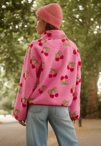Rosa Fleecejacke mit Kirschblütenmuster und grünen Akzenten, kombiniert mit hellblauen Jeans. Das Model trägt eine rosa gerippte Mütze.