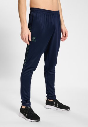Männliches Model trägt marineblaue Hummel-Trainingshose mit grünen Chevron-Streifen und schwarzen Sportschuhen, steht vor weißem Hintergrund.
