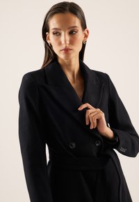 Femme aux longs cheveux foncés portant des boucles d'oreilles créoles et un manteau noir à double boutonnage avec ceinture, tenant le revers et regardant sur le côté.