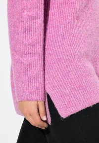 Rosa stickad tröja med ribbad textur, med en sidsprund och mjukt, något ludet material. Hand synlig som håller i tyget.