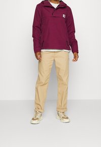 Veste anorak marron avec une poche avant, portée sur une chemise grise, associée à un pantalon beige clair et des baskets beiges.