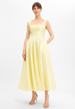 Vrouw staand in een bleekgele mouwloze midi-jurk met een vierkante halslijn en geplooide rok, met gouden sandalen met hoge hakken en bandjes.