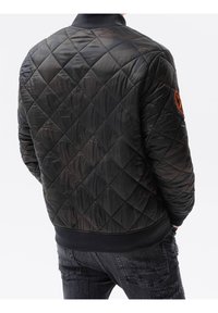 Sort quiltet bomberjakke med ribbet krave, orange emblemdetalje på ærmet og en glat, struktureret finish.