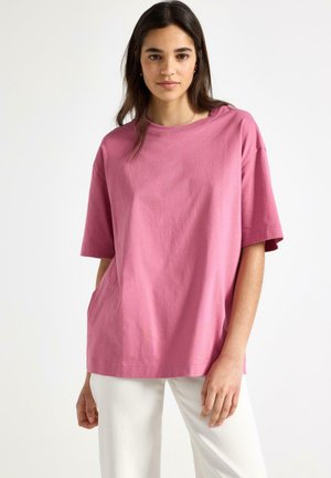 Nuori nainen, joka pukeutunut löysään vaaleanpunaiseen lyhythihaiseen paitaan ja valkoisiin housuihin, seisoo neutraali ilme kasvoillaan yksiväristä taustaa vasten.