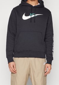 Hoodie noir avec un grand logo Nike blanc, poche kangourou et texte imprimé sur la manche droite. Tissu doux, coupe décontractée, cordons réglables.
