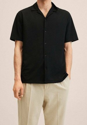 Mand iført en kortærmet sort skjorte med knapper, løst stoppet ned i beige plisserede bukser, stående med den ene hånd i lommen.