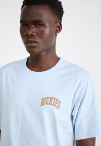 Világoskék pamut póló, kerek nyakkivágással és narancssárga "DICKIES" hímzéssel a bal mellrészen. Rövid ujjú.