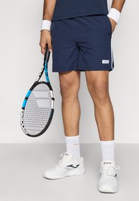 Tamsiai mėlyni teniso šortai su baltais šonų juostelėmis, derinami su baltais sportiniais batais ir tekstūruotomis baltomis kojinėmis, laikant juodą ir mėlyną teniso raketę.