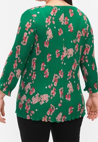 Blusa verde con motivo floreale in rosso e beige, caratterizzata da una texture plissettata e maniche a tre quarti. Dettaglio a pince sul bordo inferiore.