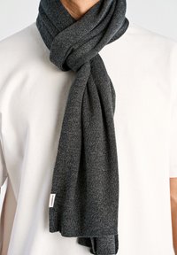 Donkergrijze gebreide sjaal, zachte textuur, om de nek gewikkeld, met een klein merketiket zichtbaar aan het uiteinde, over een wit shirt gedragen.