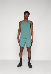 Minkštas, laisvas stiliaus teal spalvos tankinis ir šortai. Šortai turi juodus šonus. Modelis dėvi juodus sportinius batelius ir kojines.