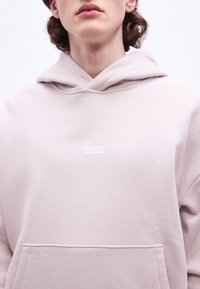 Jeune personne portant un sweat à capuche rose clair avec une poche avant et un petit logo blanc "HUGO" sur la poitrine, sur un fond blanc uni.